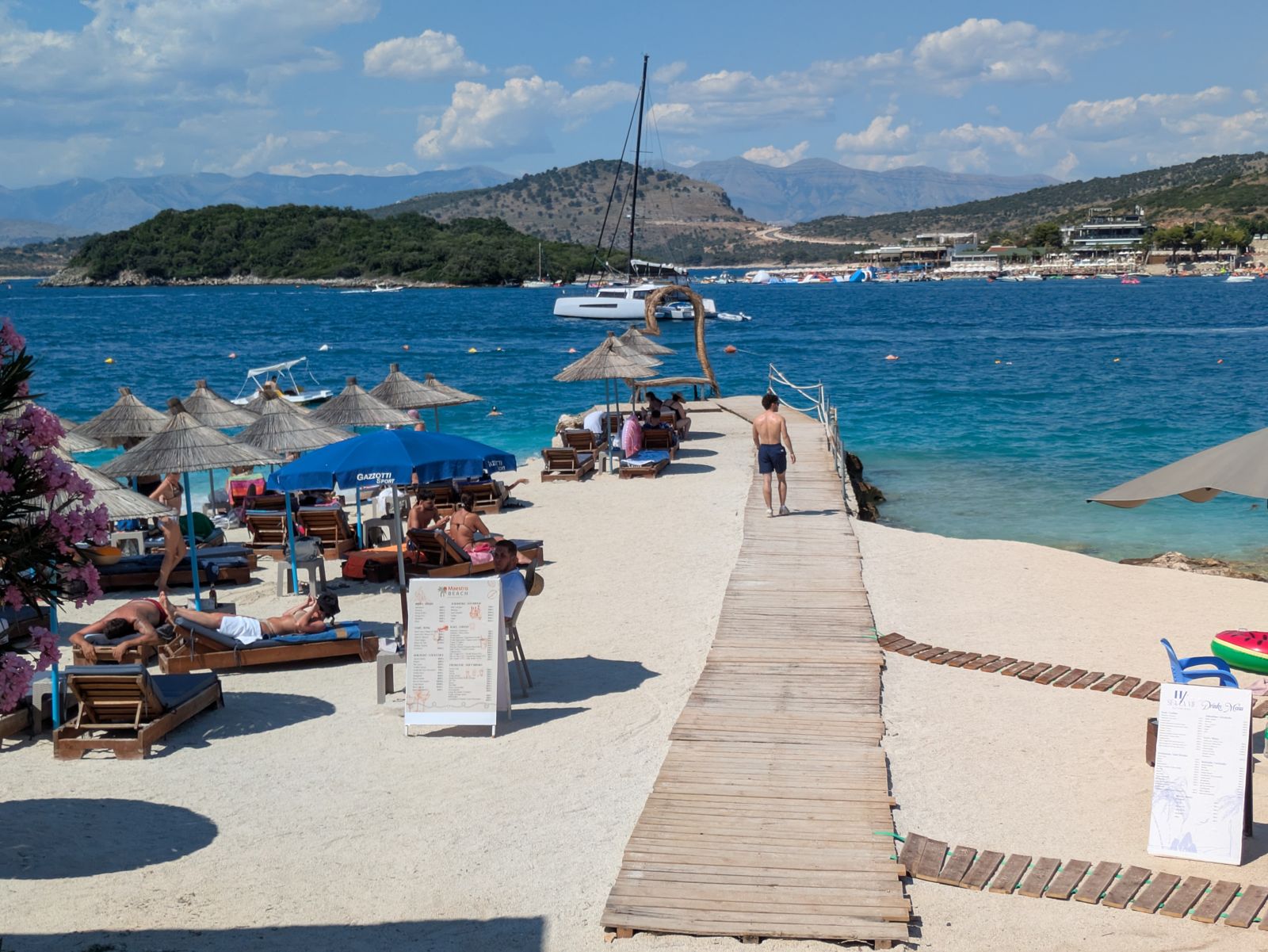 Pasarela de madera sobre la arena de una playa de Ksamil con sombrillas y veleros